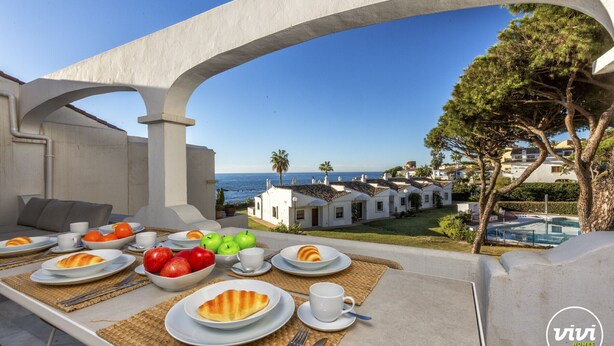 An outdoor dining area with a table set for breakfast, overlooking a coastal view with villas and a pool. - Nico | Sea Siew - Pool - Beach (Sitio de Calahonda)