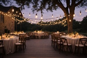 An outdoor dining area with round tables set up under string lights, surrounded by trees and a stone building.