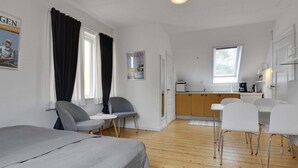 Una habitación moderna con cama, dos sillas, una mesita, kitchenette y ventana.