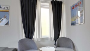 Una habitación con dos sillones tapizados de gris, una pequeña mesa blanca y una ventana con cortinas negras.