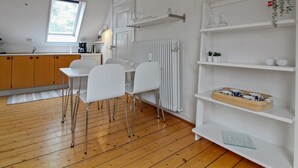 Cocina moderna con armarios de madera, encimera blanca y ventana. Se ven dos sillas blancas y una mesa blanca. En la pared, hay un radiador blanco, y una estantería con cristalería y una planta.