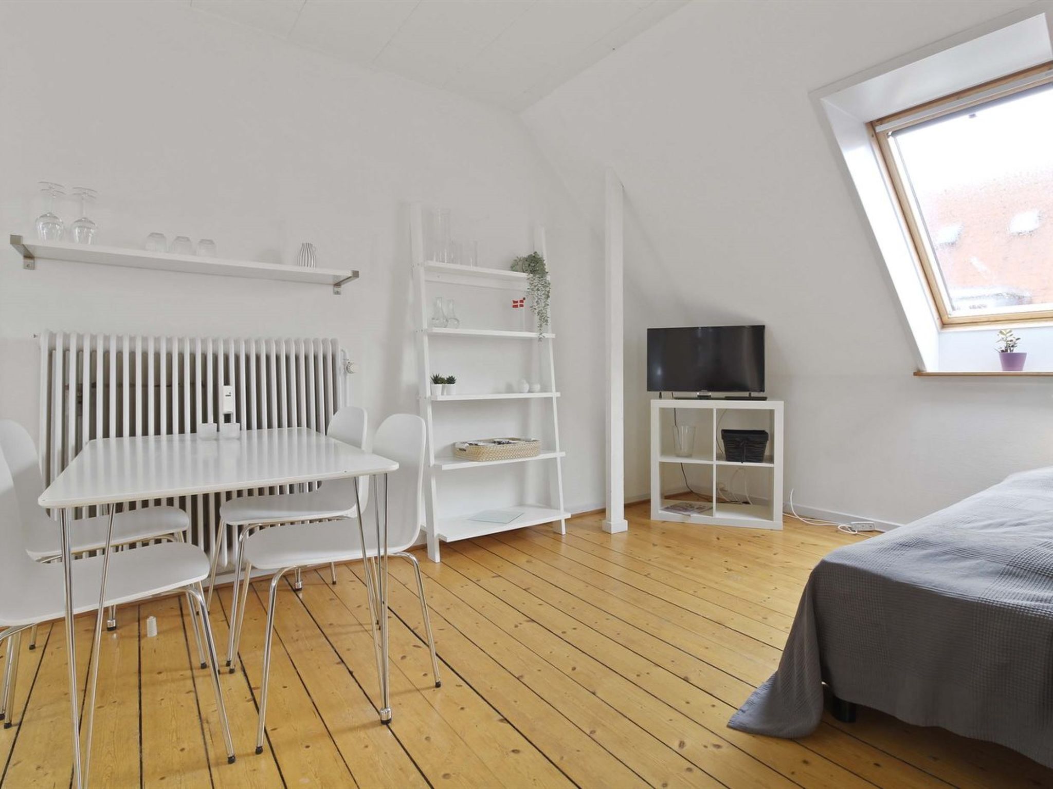 Une pièce avec un radiateur blanc, une table blanche avec des chaises, un téléviseur posé sur un meuble et une fenêtre donnant sur des immeubles.