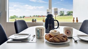 Una mesa de desayuno con pasteles, una cafetera y tazas junto a una ventana que ofrece vistas a un campo verde y edificios.