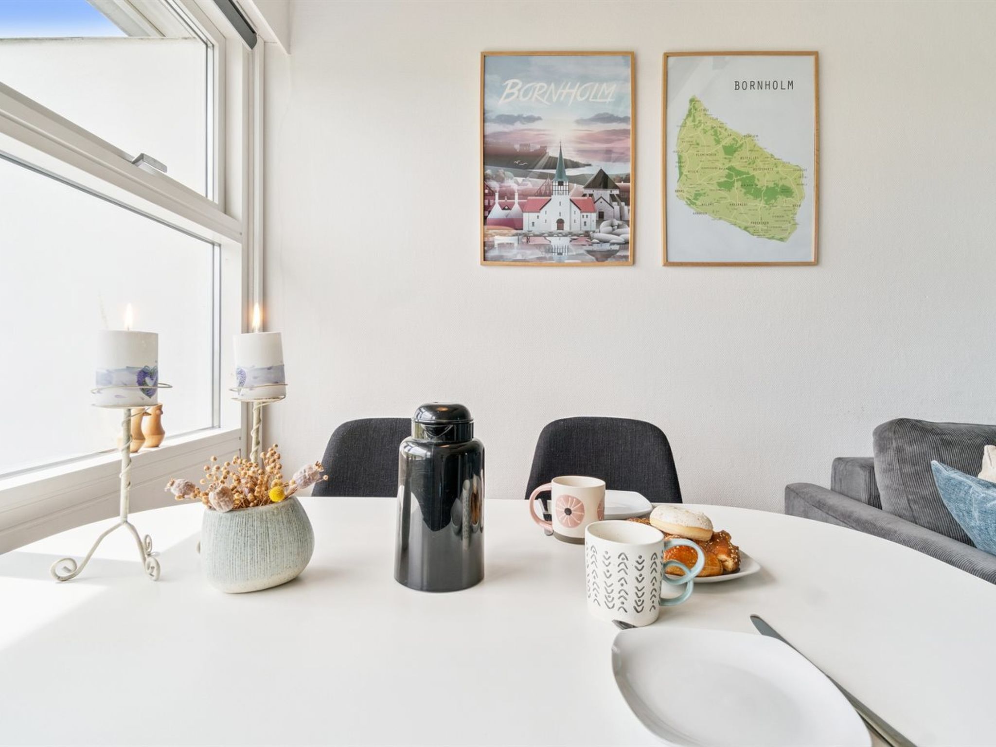 Una mesa de comedor con una vela, un termo y un plato con pasteles. Hay dos sillas y un sofá al fondo. En la pared, dos cuadros de Bornholm.
