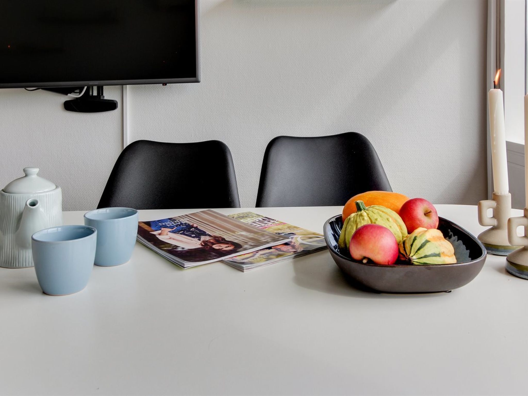 Un comedor con una mesa blanca, dos sillas negras, un cuenco de fruta, una tetera, dos tazas azules y una pila de revistas.