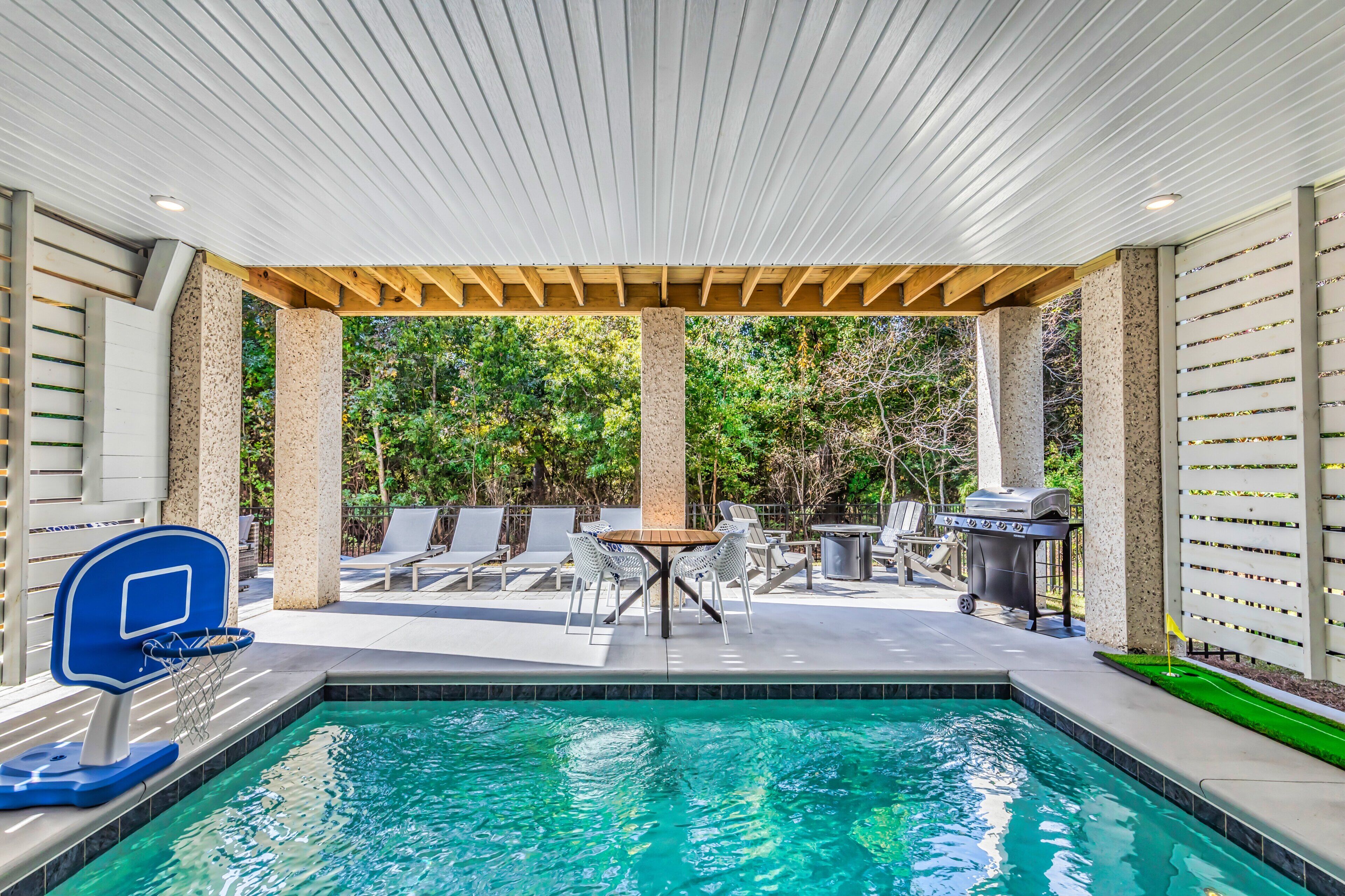 A pool area with a basketball hoop, a table with chairs, and a grill.