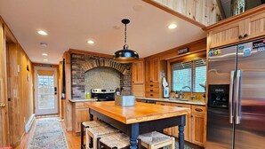 A kitchen with wooden cabinets, a stone fireplace, a stainless steel refrigerator, and a wooden dining table with bar stools. - Holocene Chalet - treetop mountain views! (Basye)