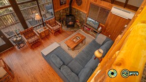 A cozy cabin interior with a stone fireplace, wooden furniture, and a large window overlooking a forest. - Holocene Chalet - treetop mountain views! (Basye)
