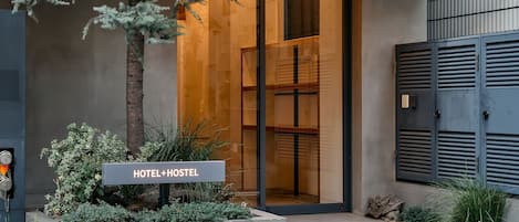 A hotel entrance with a sign reading "HOTEL+HOSTEL," a concrete planter with greenery, and a modern building facade.