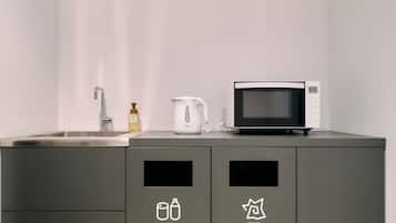 Eine Küche mit Spüle, Wasserkocher und Mikrowelle auf einem grauen Schrank. Es befinden sich zwei Recyclingbehälter mit den Aufschriften 'Recyclables' und 'Burnables' dort.