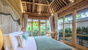 A spacious bedroom with a large bed, wooden ceiling beams, and a view of lush greenery outside. - Kaju Bungalow by Betterplace (Canggu)