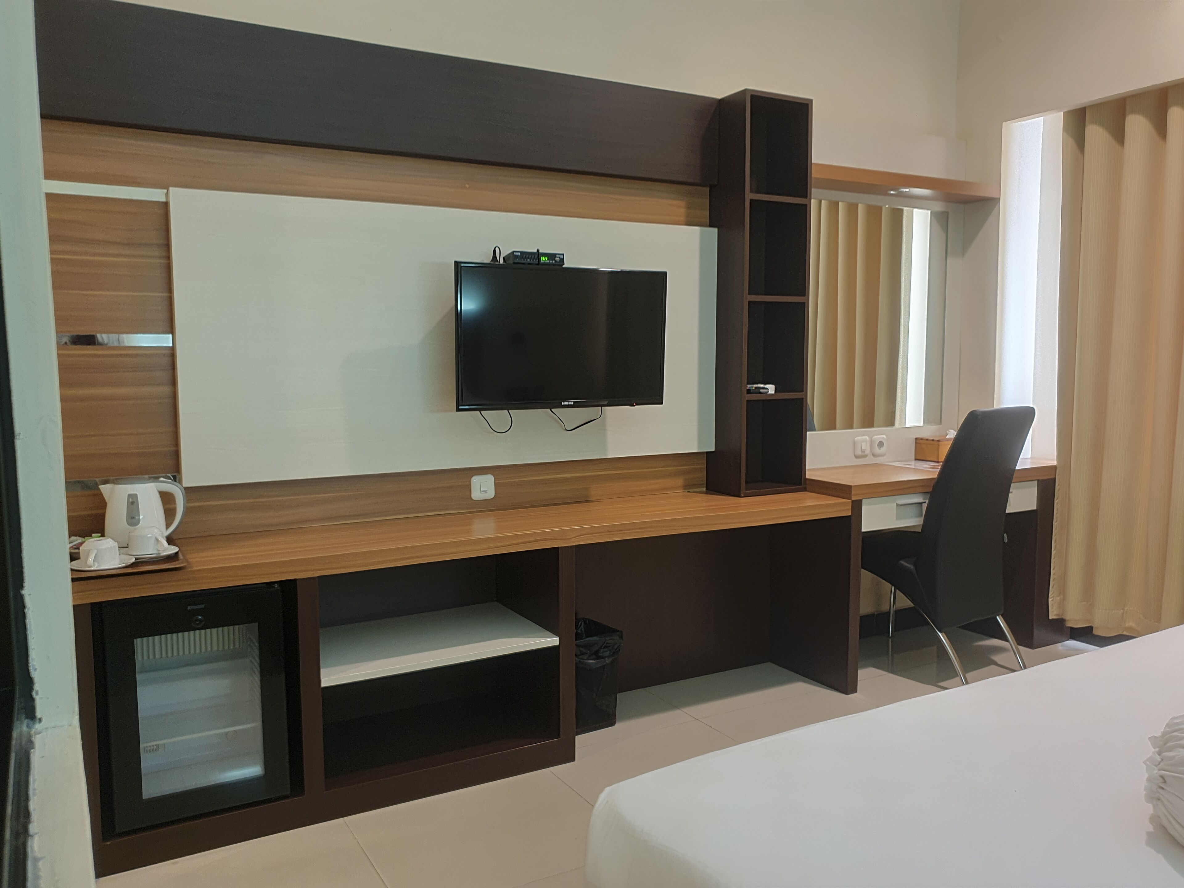 Hotelkamer met een bed, een bureau met een computer, een televisie, een minibar en een raam met gordijnen.