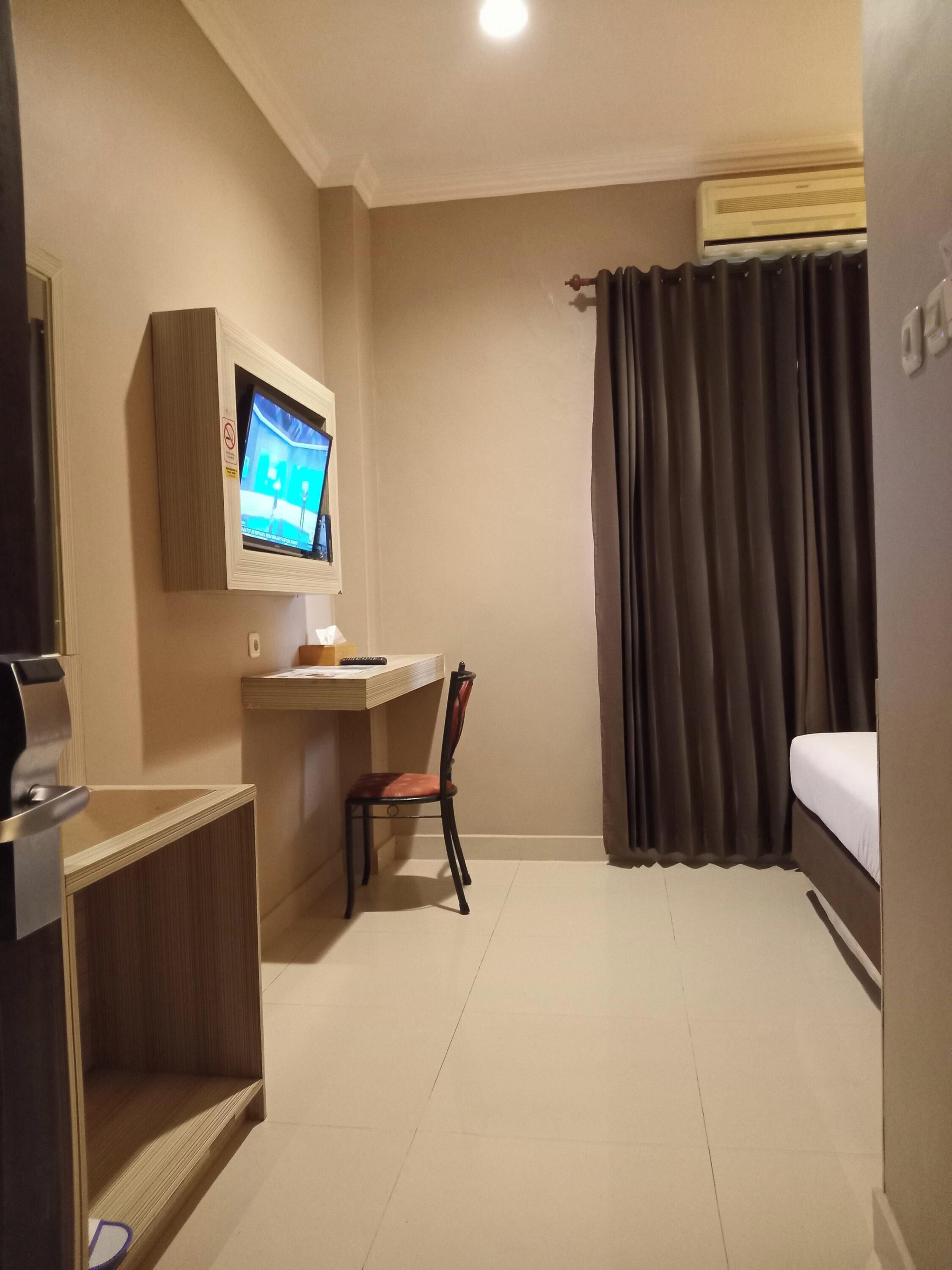 A hotel room with a flat-screen TV mounted on the wall, a desk with a chair, a bed, and a window with curtains.