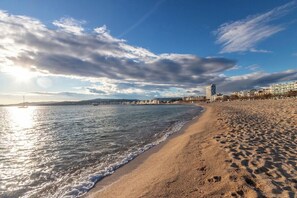 Uma praia com céu claro e mar calmo, areal arenoso e edifícios ao fundo.