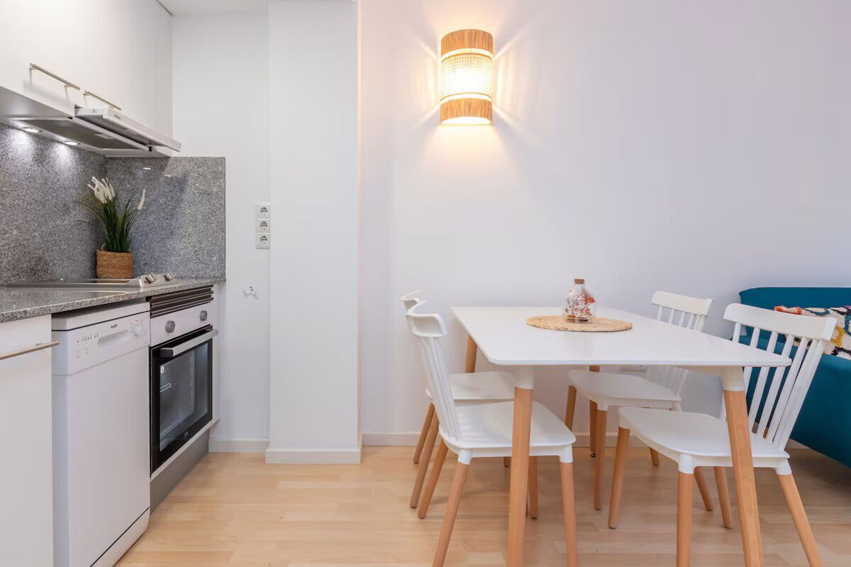 A modern kitchen with white cabinetry, a built-in oven, and a granite countertop. There is a dining area with a white table and chairs, and a wall-mounted light fixture.