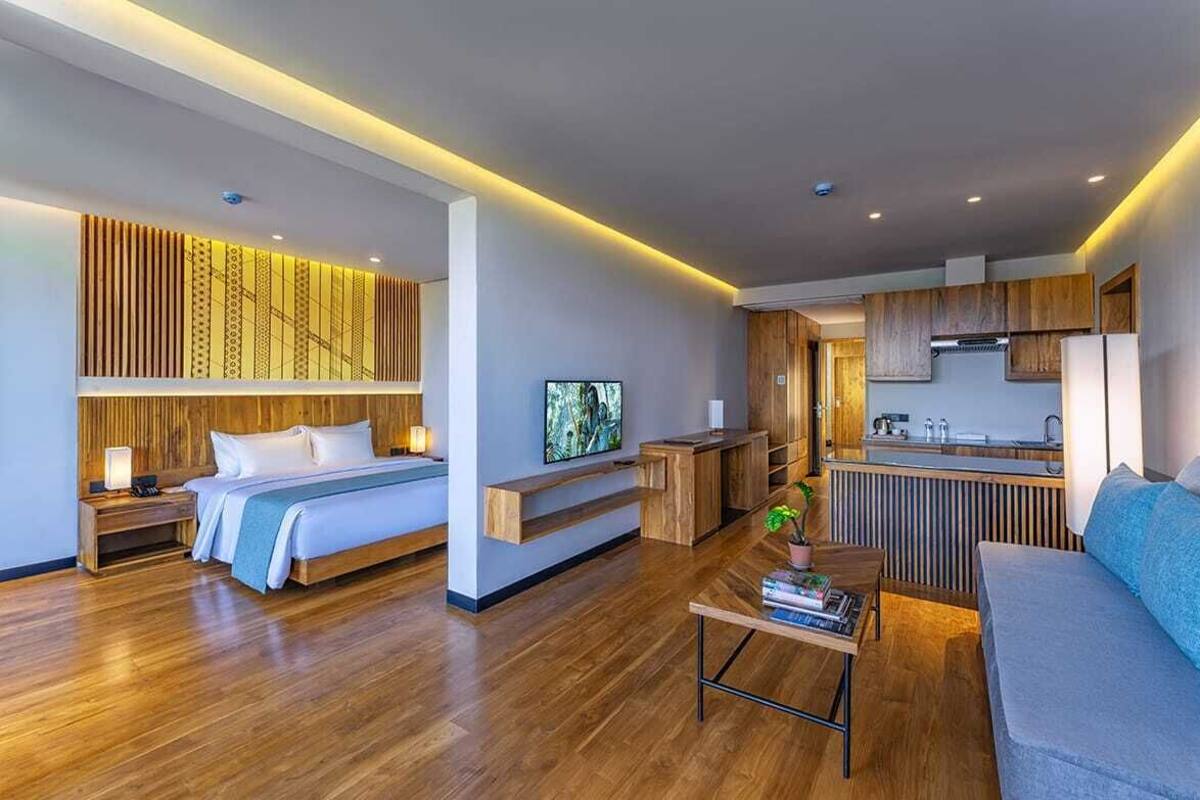 A modern hotel room with a wooden floor, a bed, a sofa, a coffee table, a TV, and a kitchenette.