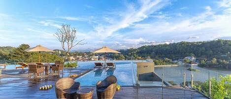 An infinity pool with wicker furniture and umbrellas, overlooking a body of water and hills.