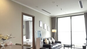 A modern living room with a dining area, a water dispenser, a sofa with cushions, and a large window providing natural light. - Vinhomes Gardenia - 2 Brs Apartment (Hanoi)