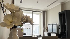 A modern living room with a large window, a white vase with flowers, a flat-screen TV, and a dark cabinet. - Vinhomes Gardenia - 2 Brs Apartment (Hanoi)