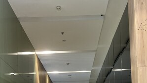 A hallway with a person walking, glass walls, a wooden panel wall with mailboxes, and a ceiling with recessed lighting. - Vinhomes Gardenia - 2 Brs Apartment (Hanoi)