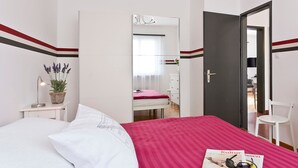 Un dormitorio con cama, una mesita de noche con lámpara y planta, un espejo, una puerta y una revista sobre la cama.