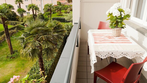 Un balcón con una mesa, una silla, una planta en maceta y vistas a un jardín y edificios.