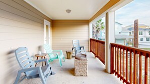 A covered porch with light blue plastic chairs, a small table, and a woven stool. - Shared Heated Pool, Golf Cart Access, Parking for 2, Pet Friendly (Port Aransas)