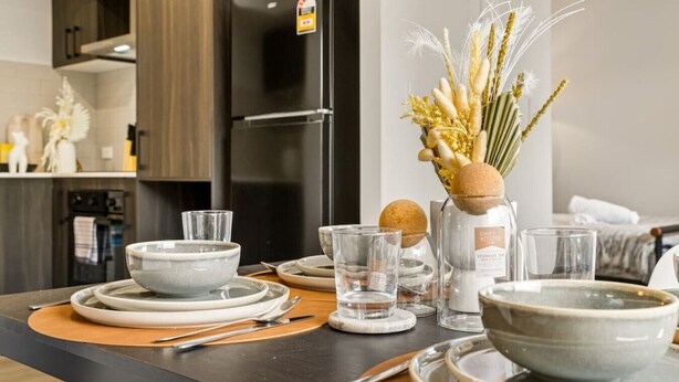 Une cuisine moderne avec un réfrigérateur noir, une table à manger dressée avec des assiettes, des bols et des verres, et un vase contenant des plantes séchées.