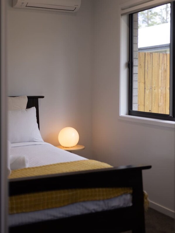 A bedroom with a bed, a bedside table with a lamp, and a window with a view of a wooden fence.