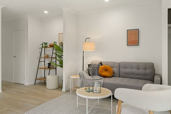 A modern living room with a grey sofa, a white armchair, a small round table, and a floor lamp. There is a ladder with shelves against the wall and a door leading to another room. - Dual-key 5BR Family home w/ stunning interior (Browns Plains)