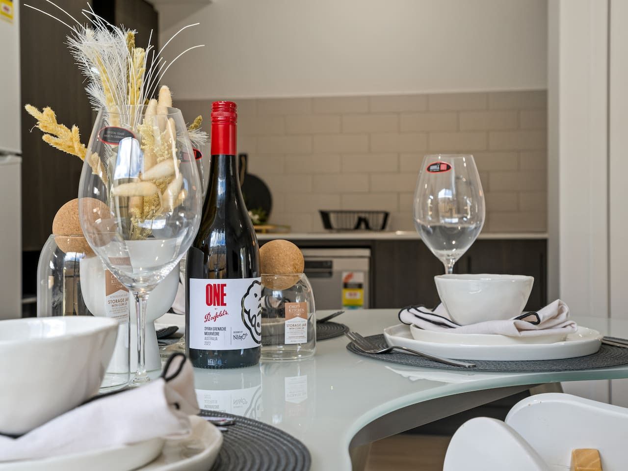 Une table à manger dressée avec des verres à vin, une bouteille de vin et un vase décoratif contenant des plantes séchées.