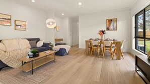 Una sala de estar moderna con sofá, una mesa de centro, una mesa de comedor y sillas. Hay cuadros en las paredes y un ventanal que ofrece vistas al exterior.