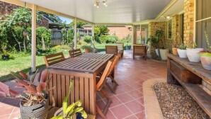 Un patio al aire libre con una mesa y sillas de madera, rodeado de plantas en macetas y un muro de piedra.