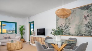 Ein modernes Wohnzimmer mit einem Esstisch aus Glas, grauen Polsterstühlen, einer gewebten Pendelleuchte, einem Flachbildfernseher und einem großen Wandbild mit tropischem Motiv.