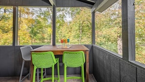 Un área de estar techada al aire libre con una mesa y dos sillas, con vistas a un bosque.