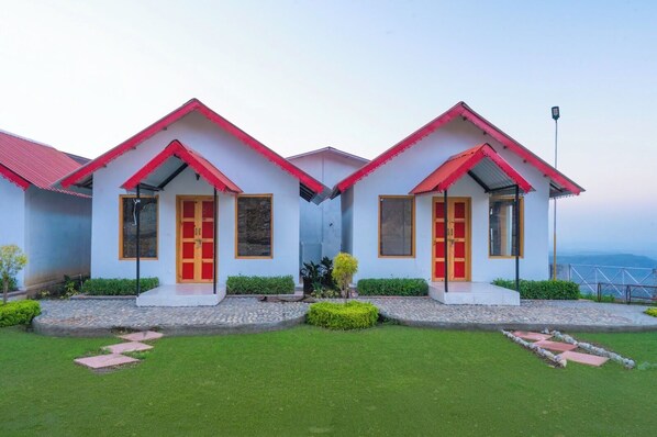 A row of two-story buildings with red roofs and white walls, featuring large windows and doors, surrounded by well-maintained lawns and pathways. - Paatli Fort Estate Resort (RAMNAGAR)