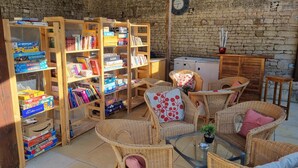 Een gezellige kamer met rieten meubels, houten boekenplanken vol boeken en spelletjes, een kleine tafel met een plant en een klok aan de muur.