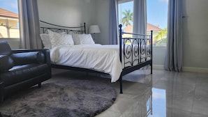 Ein Schlafzimmer mit einem Bett, einer schwarzen Ledersofa, einem flauschigen Teppich und Blick auf Palmen durch große Fenster.