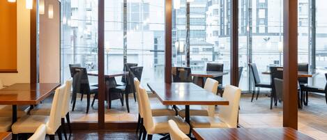 A modern dining area with wooden tables and white chairs, large windows offering a view of buildings outside.