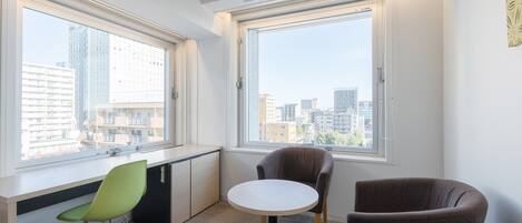 Una habitación moderna y luminosa con ventanas amplias que ofrecen vistas de la ciudad, un escritorio con silla, y una mesa redonda con dos sillas.