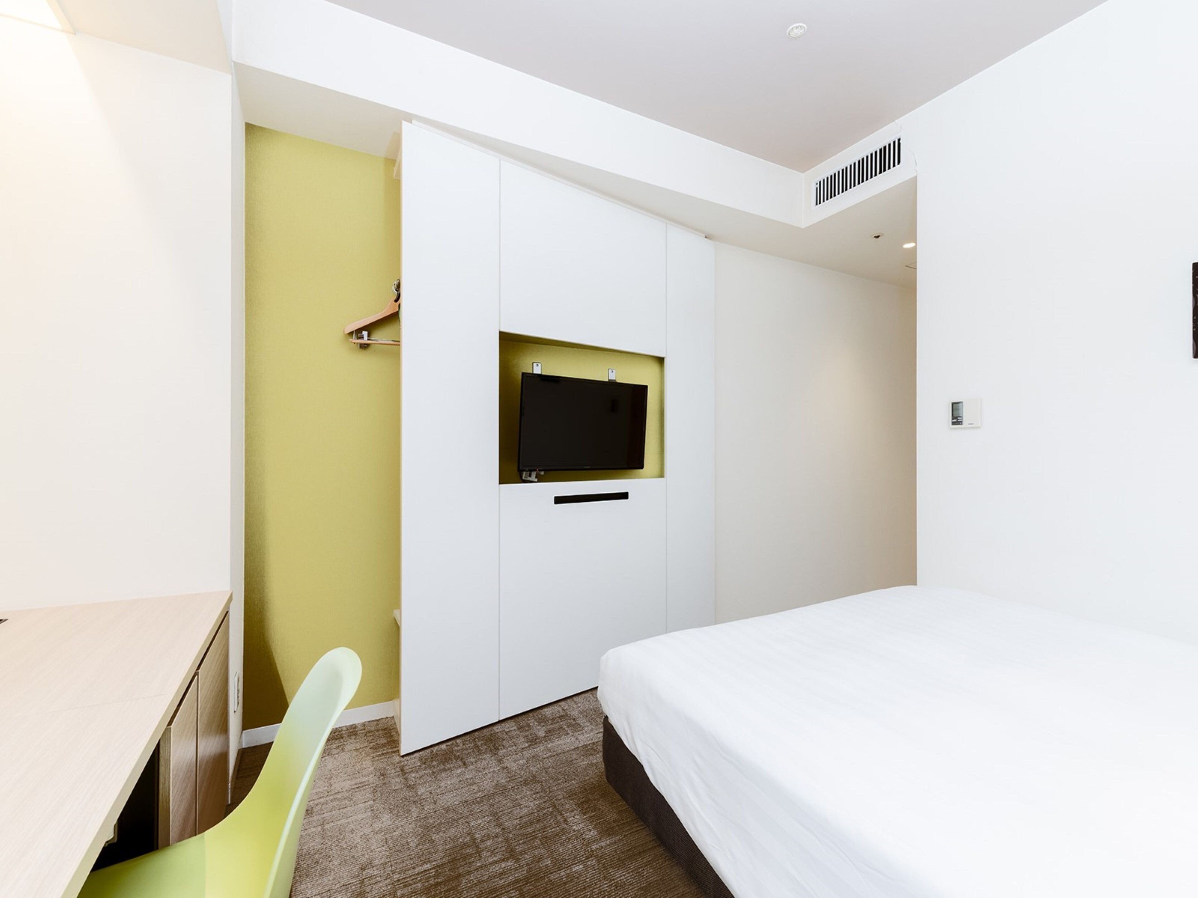 A hotel room with a bed, a desk, a chair, a TV mounted on a white wardrobe, and a small window.
