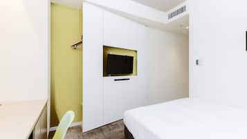 Habitación de hotel con una cama, un escritorio, una silla, un televisor montado en un armario blanco y un ventanal pequeño.