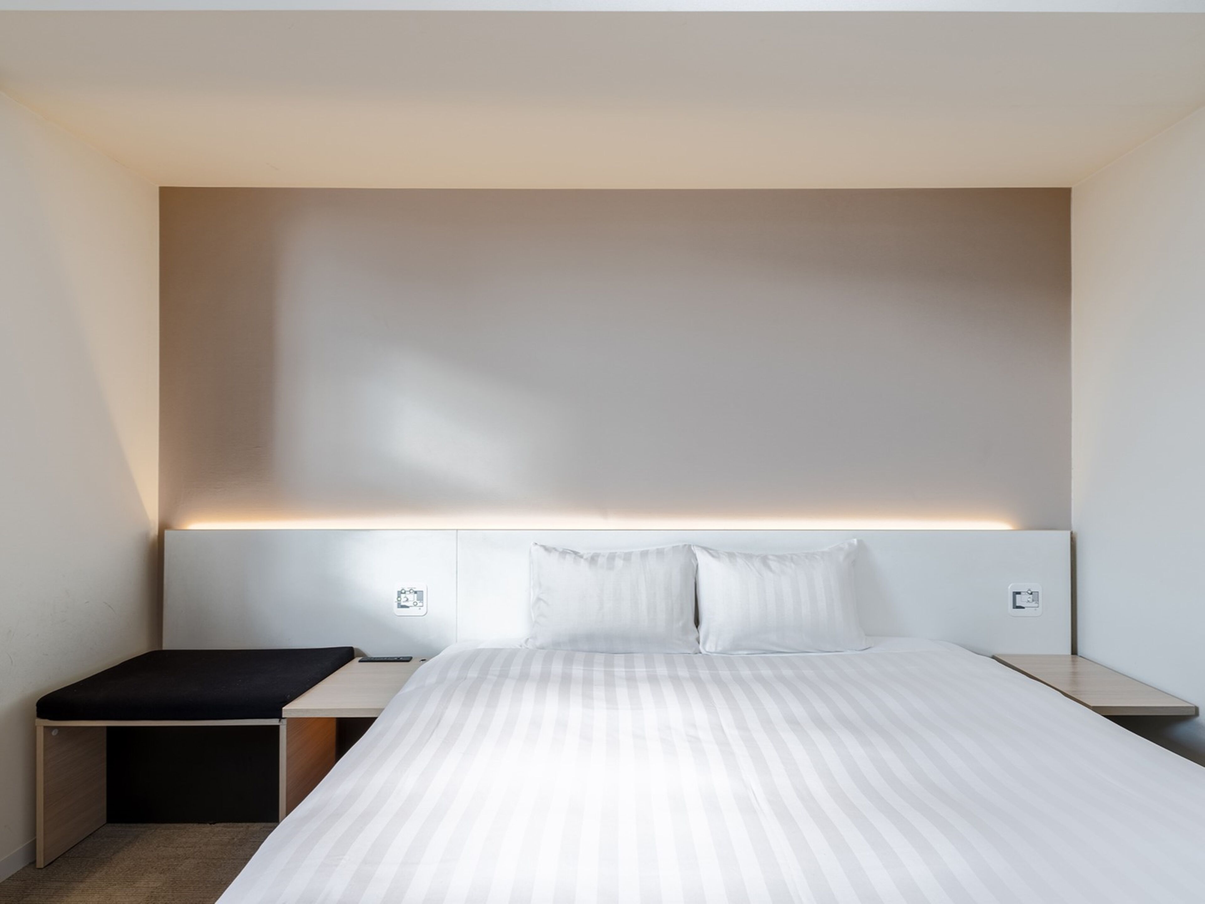 A modern hotel room with a large bed, a bedside table, a small bench, and a wall-mounted light fixture.