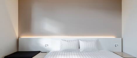 A modern hotel room with a large bed, a bedside table, a small bench, and a wall-mounted light fixture.