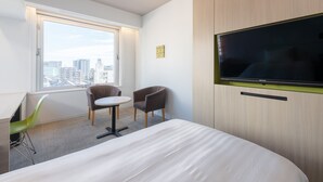 Une chambre d’hôtel moderne avec un grand lit, un téléviseur fixé au mur, une petite table ronde, deux chaises et une fenêtre donnant sur la ville.