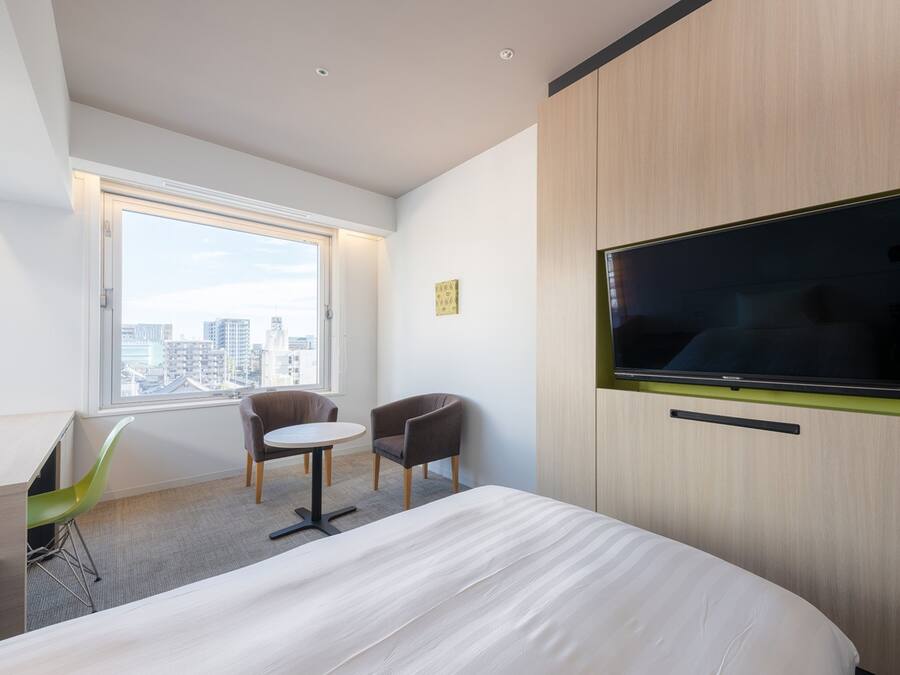 Une chambre d’hôtel moderne avec un grand lit, un téléviseur fixé au mur, une petite table ronde, deux chaises et une fenêtre donnant sur la ville.