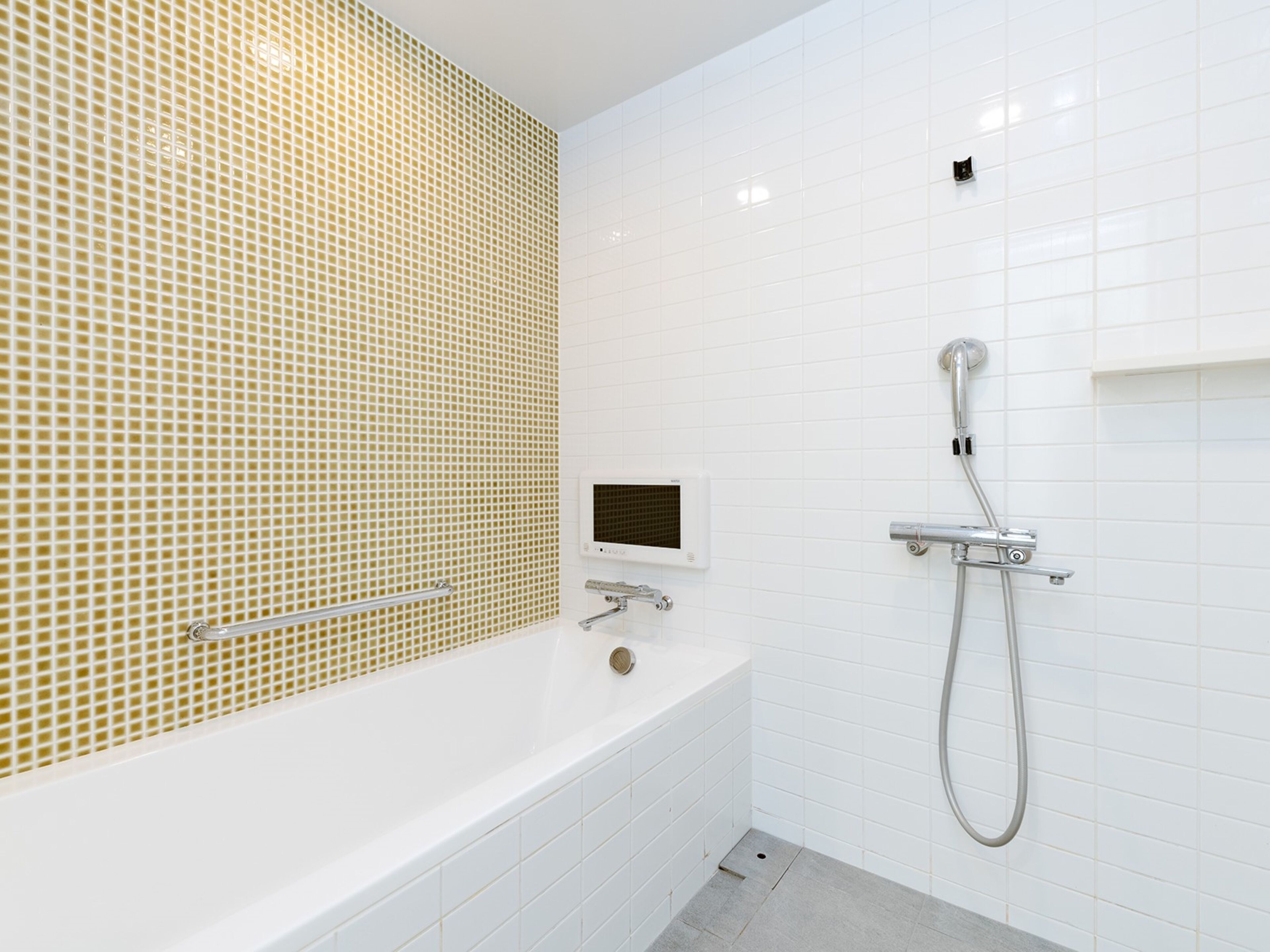 Une salle de bain moderne avec un mur recouvert de carreaux blancs, un pare-baignoire en mosaïque beige, une baignoire blanche et une douche équipée d’un pommeau de douche à main.