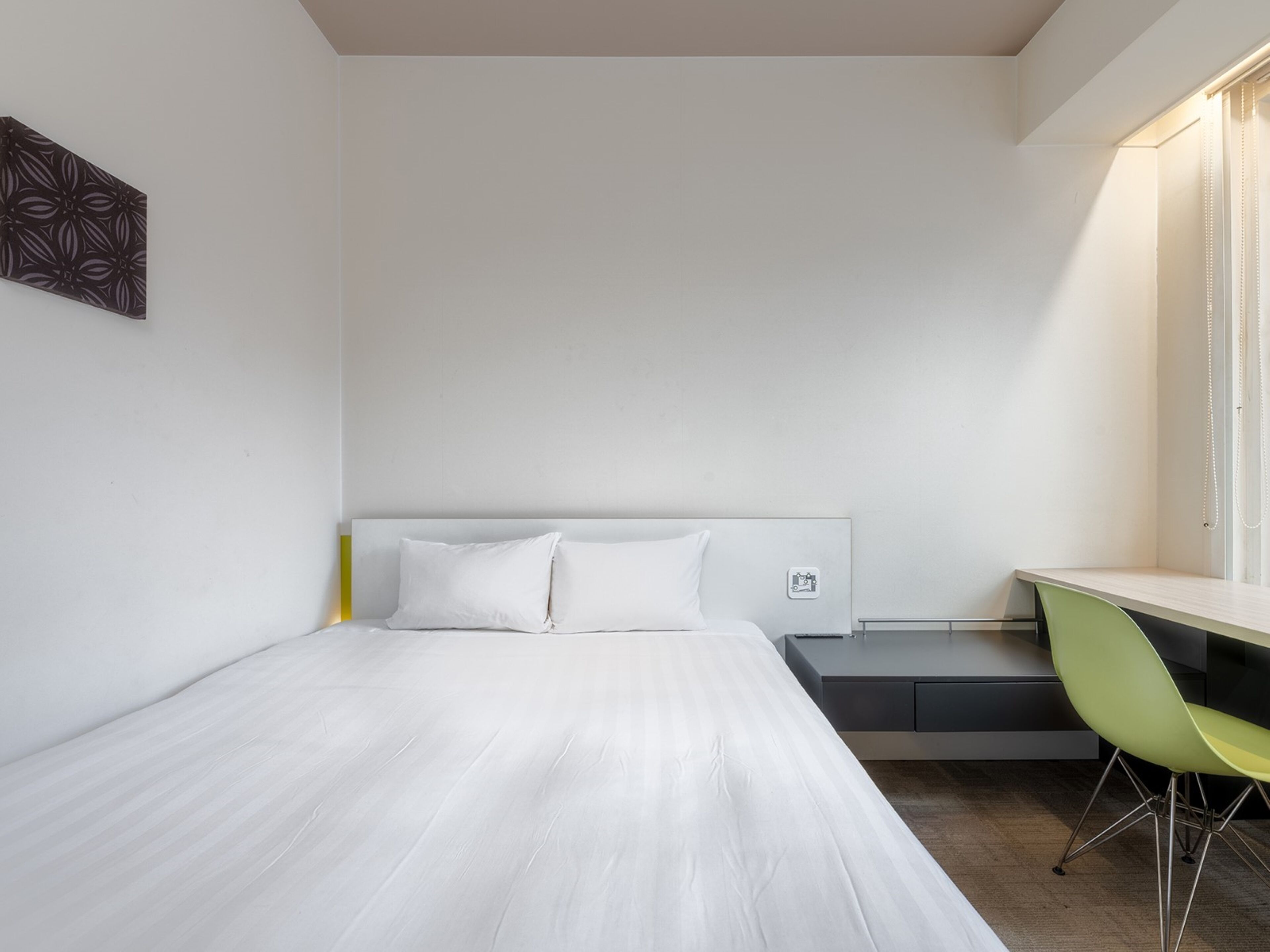 A minimalist hotel room with a bed, a desk, and a green chair.