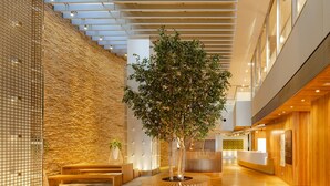 Un hall d’entrée moderne avec un mur en pierre, des bancs en bois, un arbre central et un comptoir d’accueil.