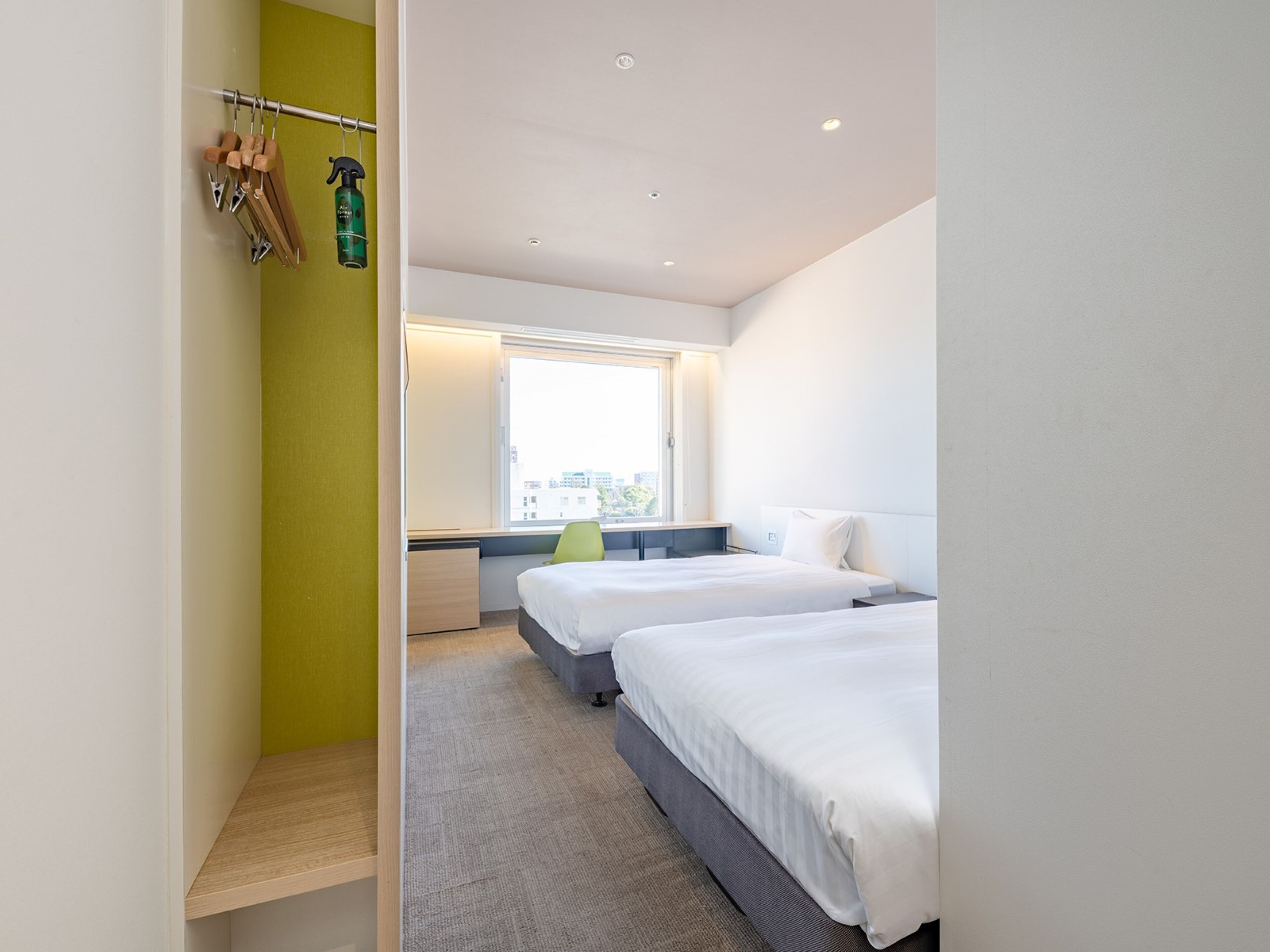 A modern hotel room with two beds, a window with a city view, a desk, and a green wall.